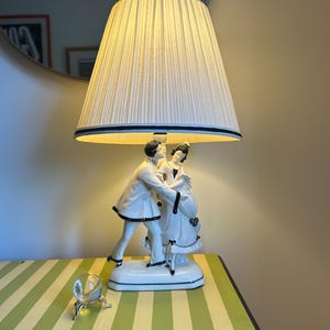 Peut inclure: Une lampe de table vintage avec un abat-jour plissé crème, bordé de bandes noires et bleues. La base présente une figurine en porcelaine blanche d'un couple dansant. La lampe est posée sur une table à rayures vertes et jaunes.