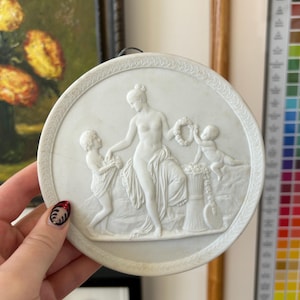 May include: A white, round bas-relief plaque depicting a classical scene with a central female figure and two cherubic figures. The figures are draped in fabric, and the plaque has a decorative border. The artwork is likely made of porcelain or a similar material.