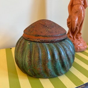 May include: A round, lidded container with a ribbed, green and brown body and a reddish-brown top. The container sits on a yellow and green striped surface. A terracotta-coloured statue is in the background.