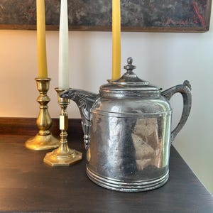 May include: A silver teapot with a decorative handle and lid sits on a dark wooden surface. Two yellow and one white taper candles stand in brass candlesticks to the left of the teapot. A painting hangs in the background.