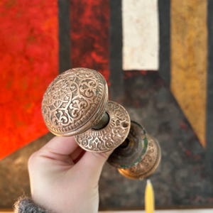 May include: Ornate antique door knobs with detailed floral patterns. The knobs are made of a tarnished metal, showing signs of age and use. The design includes a round knob and a circular backplate. The background features a colorful abstract painting.