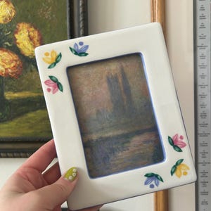 May include: White rectangular picture frame with a floral design in yellow, pink, and blue. The frame holds a landscape painting with muted colors. The frame is held in a hand.