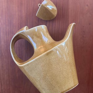 May include: A mustard-colored ceramic teapot with a distinct crackle glaze finish, featuring an integrated handle and a long, tapered spout. The teapot includes a separate, matching triangular lid with a small knob, showcasing a uniform texture.
