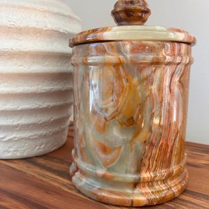May include: A cylindrical stone canister featuring a polished finish with swirling patterns of orange, brown, cream, and translucent green. The container includes a matching removable lid with a rounded knob handle, showcasing natural mineral banding.