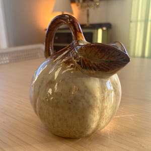 May include: A ceramic pitcher with a pumpkin-like shape. The pitcher is primarily beige with brown accents on the handle and leaf-shaped spout. The handle curves gracefully, and the spout is detailed with a leaf design. The pitcher sits on a wooden surface.