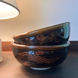 May include: Two ceramic bowls stacked on top of each other. The bowls are dark brown with a glossy finish and a wave-like pattern in a lighter brown color. The bowls are sitting on a dark brown surface.