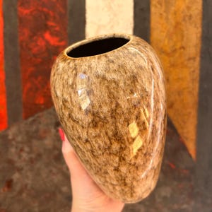 May include: A ceramic vase with a rounded, bulbous shape and a smooth, glossy finish. The surface features a mottled, speckled pattern in shades of brown, tan, and cream, creating a textured appearance. The interior of the vase is dark.