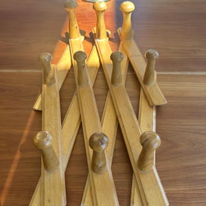 May include: A light-colored wooden accordion-style coat rack with multiple pegs. The rack is made of thin, rectangular wooden slats and has a series of rounded pegs for hanging items. The rack is designed to expand and contract, and is shown on a wooden surface.