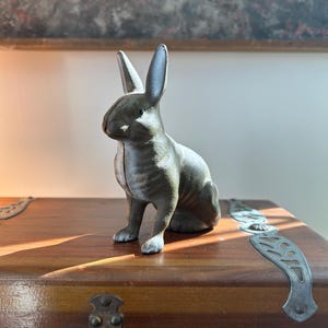 May include: A gray cast iron rabbit figurine with long ears, sitting upright. The bunny is on a wooden trunk with metal accents. The background includes a painting with red and gray tones.