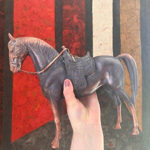 May include: A bronze-colored horse figurine with a saddle and bridle. The horse is held in front of a colorful background with red, black, and tan vertical stripes. The figurine has detailed features and a metallic finish.
