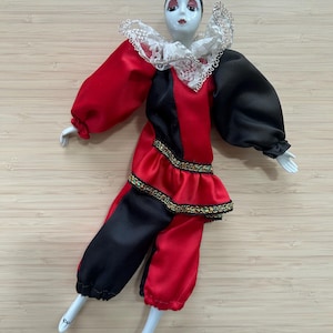 Peut inclure: Poupée en porcelaine vêtue d'un costume d'arlequin. La tenue de la poupée est en satin rouge et noir avec une jupe à volants et un col en dentelle blanche. La poupée a un visage peint avec des lèvres rouges et un bonnet noir.