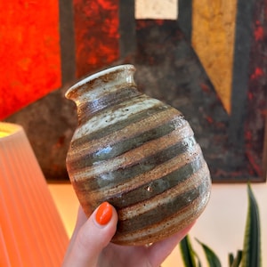 May include: A small, bulbous ceramic vase featuring a textured surface with horizontal bands of earthy brown, muted green, and cream tones. The vessel has a narrow, flared rim and a rounded body, showcasing a hand-thrown, rustic aesthetic.