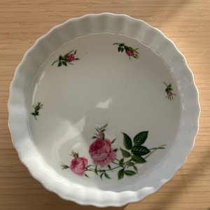 May include: White ceramic pie dish with a scalloped edge. The dish is decorated with pink roses and green leaves. The pie dish is round and sits on a wooden surface.