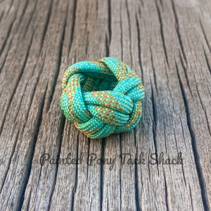 May include: A decorative knot made of turquoise, green, yellow, and orange rope. The knot is centered on a weathered wooden surface. The text "Painted Pony Tack Shack" is at the bottom of the image.