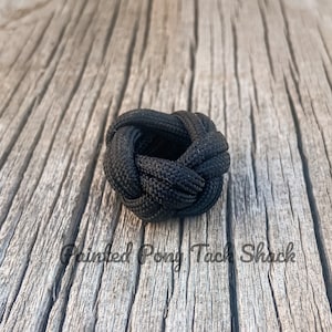 May include: A black paracord ring knot, also known as a cobra knot, is tied in a circular shape. The knot is made with a thick black cord and is sitting on a wooden surface.
