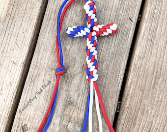 May include: A red, white, and blue paracord cross keychain. The keychain is made of braided paracord and has a loop at the top for attaching to keys or a bag.