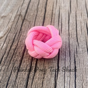 May include: A pink paracord knot, also known as a cobra knot, is tied in a circular shape. The knot is made with a thick, pink paracord and is sitting on a wooden surface.