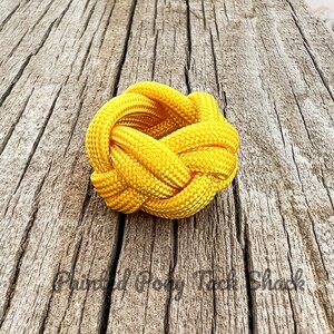 May include: A yellow paracord knot tied in a circular shape. The knot is made of thick, braided rope and is sitting on a wooden surface.