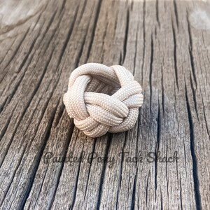 May include: A white paracord ring knot, tied in a complex pattern, is displayed on a weathered wooden surface.
