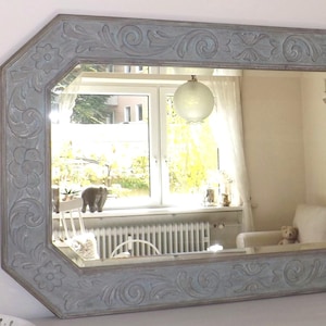 Könnte beinhalten: Rechteckiger Spiegel mit hellblauem, verziertem Rahmen. Der Rahmen weist geschnitzte Blumen- und Scrollmuster auf. Der Spiegel spiegelt einen Raum mit Fenster, Sofa und Dekorationsgegenständen wider.