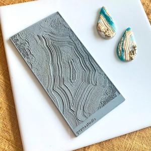 Puede incluir: Un sello rectangular gris con un patrón geológico detallado y el texto "goynastudio" se muestra en una superficie blanca. También hay dos pendientes en forma de lágrima con acentos turquesa, dorados y blancos.