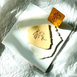 May include: A white ceramic tile with a yellow clay imprint of a floral design. The design features a branch with leaves and berries. A yellow rubber stamp with the same design is also shown.