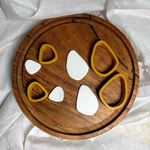 Könnte beinhalten: Ein Set mit sieben Ausstechern in verschiedenen Formen, darunter Kreise, Ovale und Tropfen. Die Ausstecher sind aus goldfarbenem Kunststoff und auf einer Holzoberfläche angeordnet.