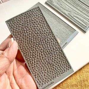 Puede incluir: Una estera de silicona gris texturizada con un patrón en relieve. La estera es rectangular y tiene el texto "goynastudio" impreso en la esquina inferior derecha.