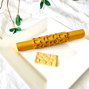 May include: A golden-yellow rolling pin with a textured, open-hole design, and a small, rectangular piece with a similar pattern. The items are on a white, rectangular plate, suggesting a baking or crafting theme.