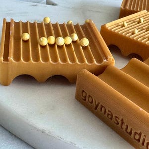 May include: Close-up of several amber-colored soap dishes with a grooved design. Each dish has a series of parallel ridges and is adorned with small, round, yellow beads. The text "goynastudio" is visible on one dish.