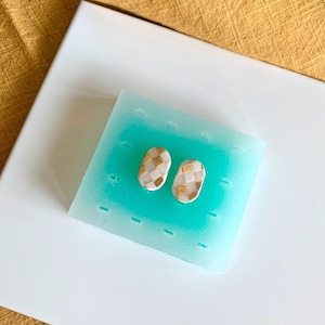 Op de afbeelding: Twee witte ovale oorbellen met een gouden geruit patroon.