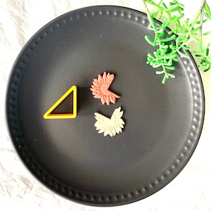 May include: Three cookie cutters on a black plate. One is a triangle shape, and the other two are floral shapes. One is red and the other is a light orange.