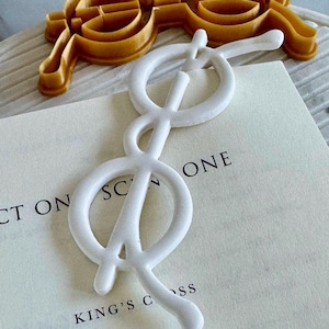 Puede incluir: Dos cortadores de galletas: uno con forma de gafas y otro con forma de nota musical. El cortador de gafas es de color marrón claro, y el de la nota musical es blanco. La nota musical está sobre un libro con el texto "CT ON SCONE" y "KING'S CSS".