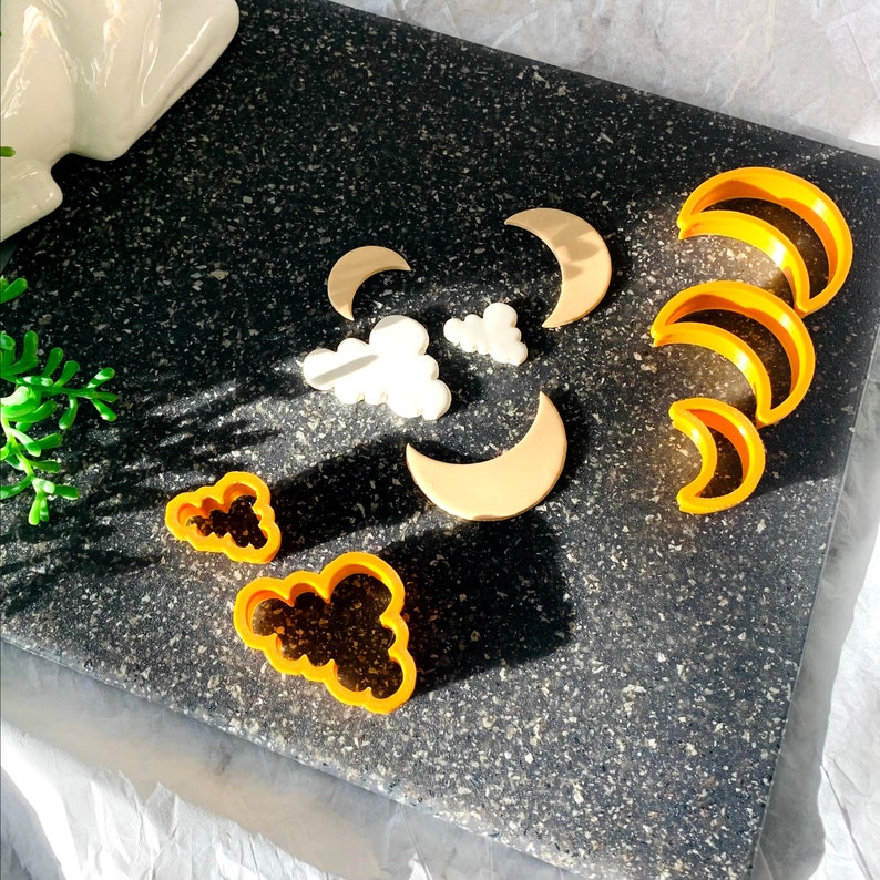 May include: A set of cookie cutters in the shape of clouds and crescent moons. The cutters are made of yellow plastic and are arranged on a black surface.