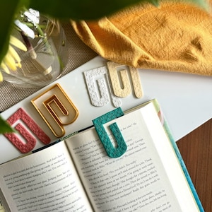 Op de afbeelding: Een close-up van een boek met een turquoise, gele en roze bladwijzer. De bladwijzer is gemaakt van geweven materiaal en heeft een uniek ontwerp. Het boek is open op een pagina met tekst over een personage genaamd Milo.