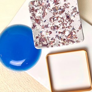 May include: Three decorative coasters, one is a round blue glass coaster, one is a square white coaster with a gold frame, and one is a square white coaster with a marbled pattern in shades of pink, brown, and white.