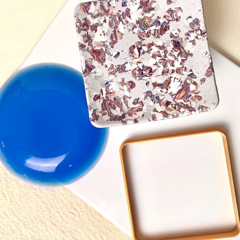 May include: Three decorative coasters, one is a round blue glass coaster, one is a square white coaster with a gold frame, and one is a square white coaster with a marbled pattern in shades of pink, brown, and white.