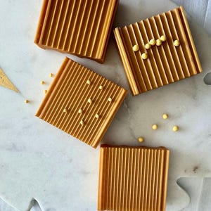 以下が含まれることがあります： 白い大理石の表面に、琥珀色の正方形の溝付き石鹸皿が4つ配置されています。 いくつかの皿には、小さな黄色の球状のアクセントがあります。 石鹸皿は暖かく魅力的な色をしています。