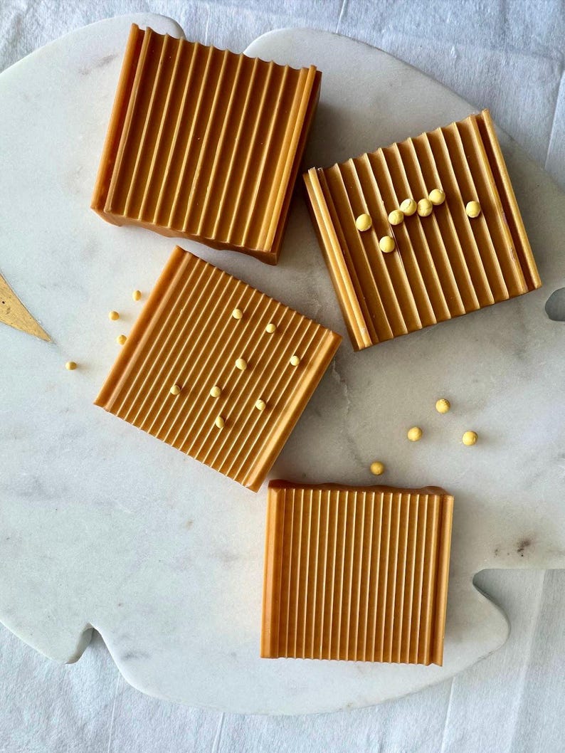 May include: Four square, ridged, amber-colored soap dishes are arranged on a white marble surface. Some dishes have small, yellow, spherical accents. The soap dishes are a warm, inviting color.
