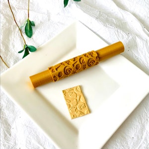 Pode incluir: Um rolo de massa de madeira com um design de conchas. O rolo está em um prato branco com um pequeno pedaço de argila que foi rolado com o rolo.