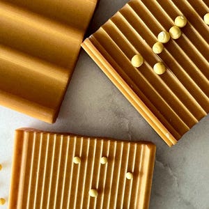 May include: Overhead shot of three rectangular, ridged, amber-colored soap dishes. Small, round, yellow objects are scattered on two of the dishes. The soap dishes are arranged on a light gray surface.