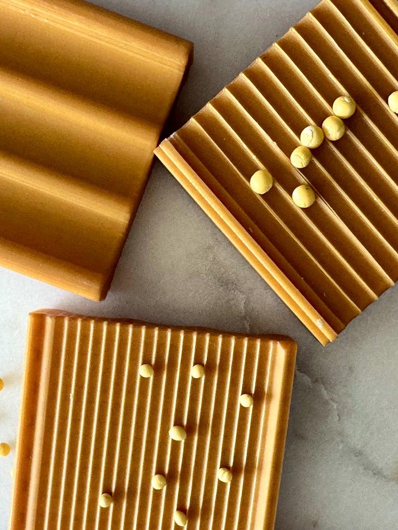 May include: Overhead shot of three rectangular, ridged, amber-colored soap dishes. Small, round, yellow objects are scattered on two of the dishes. The soap dishes are arranged on a light gray surface.