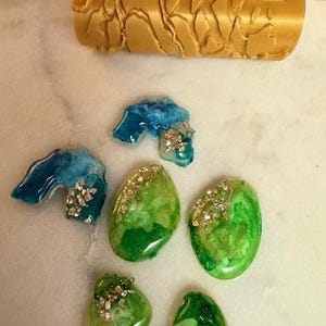 May include: A collection of handmade resin art pieces in shades of green, blue, and gold. The set includes several oval-shaped pendants with glitter accents and a textured cylindrical roller. The pieces are displayed on a marble surface.