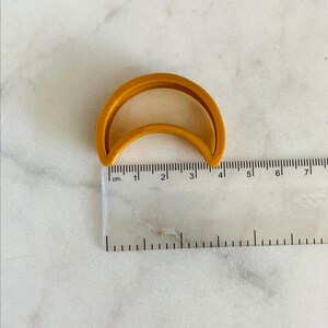 May include: A gold-colored plastic cookie cutter shaped like a crescent moon. The cutter is approximately 4 cm wide.