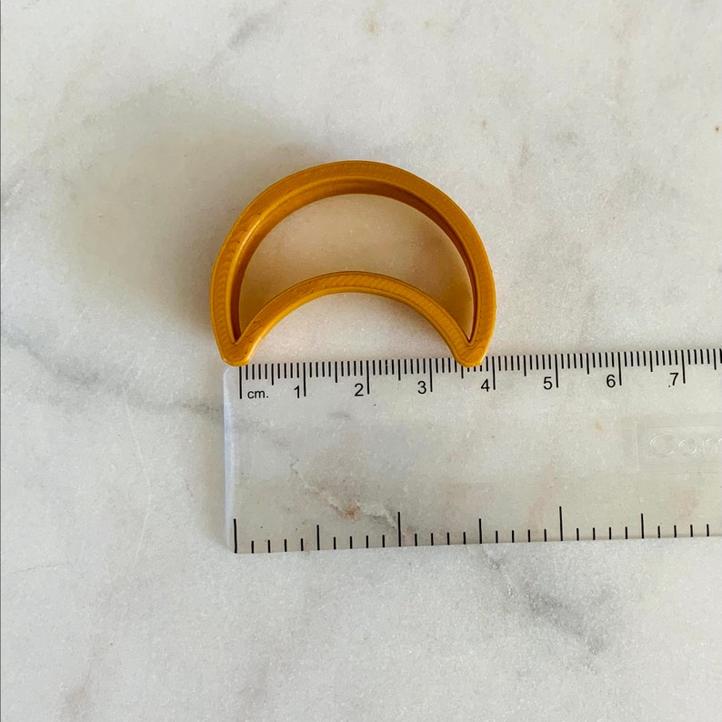 May include: A gold-colored plastic cookie cutter shaped like a crescent moon. The cutter is approximately 4 cm wide.