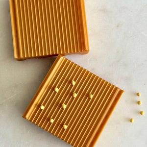 May include: Two square, amber-colored soap dishes with a ribbed design. One dish has small, yellow beads scattered across it. The dishes are on a white marble surface.