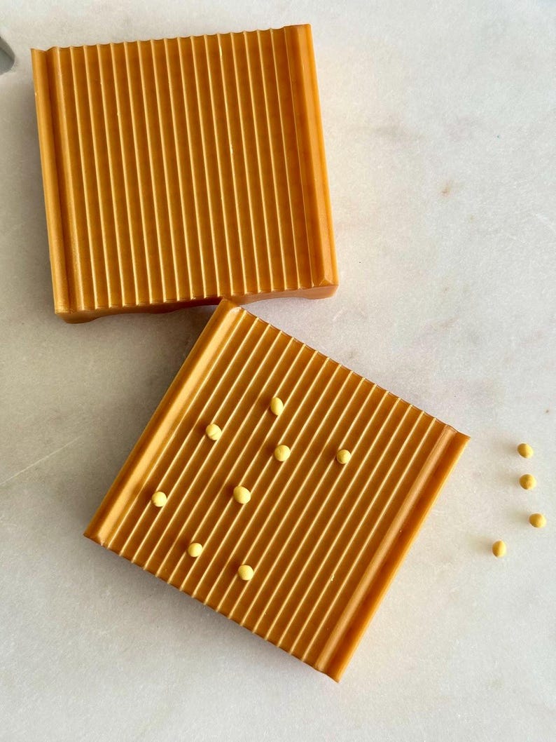 May include: Two square, amber-colored soap dishes with a ribbed design. One dish has small, yellow beads scattered across it. The dishes are on a white marble surface.