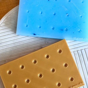 Puede incluir: Dos moldes rectangulares de silicona para manualidades. El molde superior es azul claro con 18 agujeros. El molde inferior es dorado con 18 agujeros y el texto "goynastudio" en la esquina inferior derecha.
