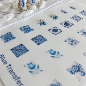 Peut inclure: Deux bagues et une feuille de motifs de transfert bleus et blancs. Les bagues présentent des motifs floraux bleus et des accents dorés. La feuille est intitulée "Blue Transfer 1" et affiche divers motifs floraux bleus.