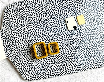 May include: Two gold-colored rectangular earrings on a white background with black polka dots.